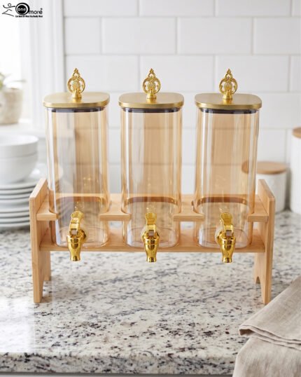 Triple 2.2L amber glass beverage dispenser set with ornate gold spigots and lids on a natural wooden stand on a marble countertop.