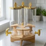 Triple glass beverage dispenser set featuring gold spigots, gold decorative lids, and a wooden rotating stand on a kitchen countertop.