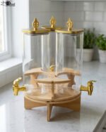 Triple glass beverage dispenser set featuring gold spigots, gold decorative lids, and a wooden rotating stand on a kitchen countertop.