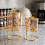 Two elegant clear glass drink dispensers with fruit prints, gold spigots, and gold deer lid handles resting on matching metal wire stands.