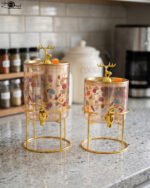 Two elegant clear glass drink dispensers with fruit prints, gold spigots, and gold deer lid handles resting on matching metal wire stands.