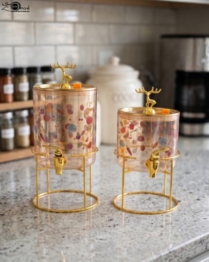 Two elegant clear glass drink dispensers with fruit prints, gold spigots, and gold deer lid handles resting on matching metal wire stands.