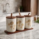 Three cream-colored ceramic kitchen storage jars featuring gold floral patterns, wooden lids with leather straps, and a matching wooden base tray sitting on a marble countertop.