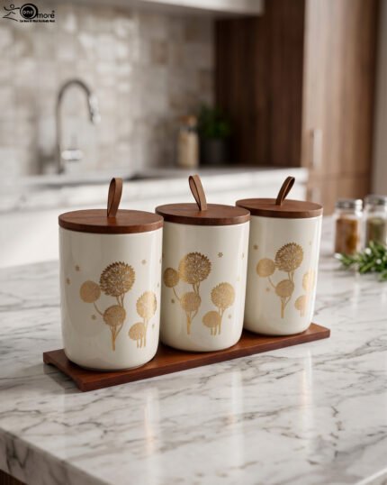 Three cream-colored ceramic kitchen storage jars featuring gold floral patterns, wooden lids with leather straps, and a matching wooden base tray sitting on a marble countertop.