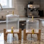 Two ribbed glass beverage dispensers with gold spigots on wooden stands sitting on a kitchen countertop