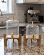 Two ribbed glass beverage dispensers with gold spigots on wooden stands sitting on a kitchen countertop