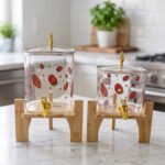 Two strawberry pattern glass drink dispensers with gold spigots set on wooden stands in a bright kitchen context.