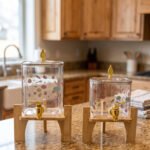 Two square glass beverage dispensers with fruit prints, gold spigots, and wooden stands on a kitchen counter