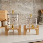Clear floral patterned glass beverage dispenser with a gold spigot set on a modern wooden stand