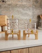 Clear floral patterned glass beverage dispenser with a gold spigot set on a modern wooden stand
