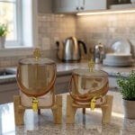 Two amber-shaded glass beverage dispensers with gold spigots, leather handles, and wooden stands on a kitchen counter.
