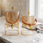 Amber shaded ribbed glass beverage dispenser with gold spigot, leather handle, and gold metal stand on a marble countertop