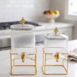 Two ribbed glass beverage dispensers with gold spigots and metal stands on a kitchen counter