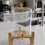 Clear 9.5L glass beverage dispenser with gold spigot on a wooden stand on a white marble countertop