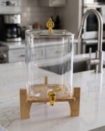 Clear 9.5L glass beverage dispenser with gold spigot on a wooden stand on a white marble countertop