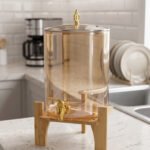 Amber glass drink dispenser with gold spigot resting on a natural wooden stand on a kitchen counter.