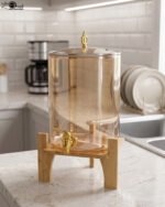 Amber glass drink dispenser with gold spigot resting on a natural wooden stand on a kitchen counter.