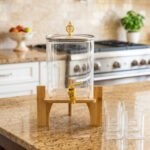 4.2L clear glass beverage dispenser with a gold spigot, gold lid handle, natural wooden stand, and 4 drinking glasses on a kitchen island.