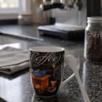 Black ceramic coffee mug printed with vintage grinder graphic and a white spoon resting in the handle slot on a granite kitchen counter.
