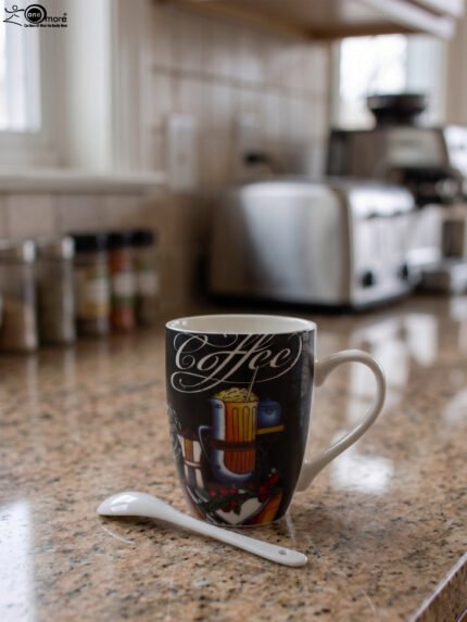 Black ceramic mug printed with vintage Coffee artwork resting on a granite kitchen counter alongside a white ceramic spoon.