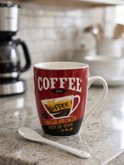 Red printed ceramic coffee mug featuring retro coffee shop logo next to a small white spoon on a kitchen counter