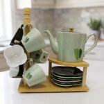 Green and black ceramic tea set with 6 cups, kettle, saucers, and a wooden display stand on a kitchen counter