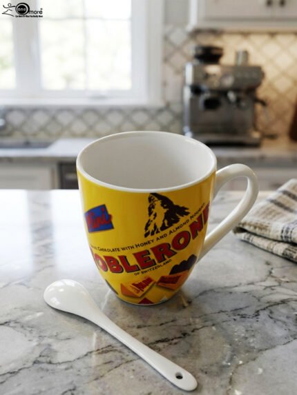 Yellow ceramic coffee mug with vintage Toblerone chocolate wrapper print and a small matching white spoon on a kitchen counter.
