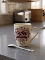 Vintage off-white ceramic coffee mug with "Coffee and then the world" text design resting on a grey countertop next to a white ceramic spoon.