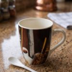 Printed ceramic coffee mug featuring coffee beans and cups artwork next to a small white ceramic spoon on a granite countertop