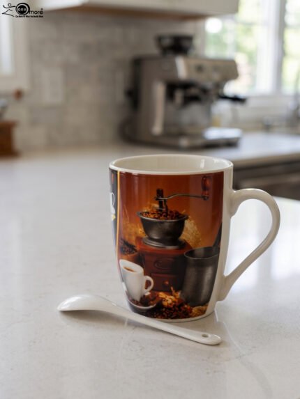White ceramic coffee mug printed with a vintage manual coffee grinder, espresso cup, and coffee beans, sitting next to a matching white spoon on a kitchen counter.