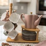 Pink and pastel ceramic tea set featuring 6 cups, 1 kettle, and a stacked saucer tier organized on a wooden display stand and tray on a kitchen counter.