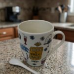 White ceramic coffee mug printed with 'Have a Nice Day' doodle art next to a matching ceramic spoon on a kitchen counter.