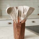 11-piece cream silicone kitchen utensil set with rose gold handles in a wooden holder on a marble countertop