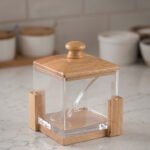 Square clear acrylic sugar pot with a natural wooden lid, clear serving spoon, and wooden base stand on a kitchen countertop.