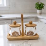 Two clear acrylic condiment jars with wooden lids and glass spoons set on a wooden stand tray with handle on a white marble countertop.