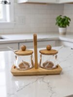 Two clear acrylic condiment jars with wooden lids and glass spoons set on a wooden stand tray with handle on a white marble countertop.