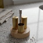 Brushed brass metal salt and pepper shakers with toothpick holder set in a round wooden base on a granite countertop