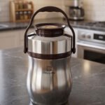 AILIJIN insulated stainless steel lunch box container with a dark brown carrying handle sitting on a modern kitchen countertop.