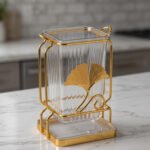 Luxury gold frame spoon stand with fluted clear acrylic container featuring a gold ginkgo leaf emblem on a marble countertop.