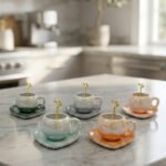 Elegant pastel ceramic cup and saucer set with a matching gold spoon and beaded handle on a marble kitchen countertop.