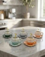 Elegant pastel ceramic cup and saucer set with a matching gold spoon and beaded handle on a marble kitchen countertop.