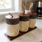 Three white and speckled black kitchen storage canisters featuring a gold deer and tree motif, fitted with wooden lids with loop handles and arranged on a matching wooden tray on a kitchen counter.