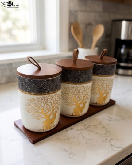 Three white and speckled black kitchen storage canisters featuring a gold deer and tree motif, fitted with wooden lids with loop handles and arranged on a matching wooden tray on a kitchen counter.
