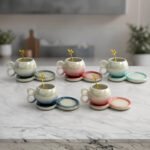 A collection of multicolored ceramic coffee cups and saucers with gold leaf spoons and double-loop handles arranged on a marble kitchen counter.