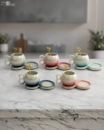 A collection of multicolored ceramic coffee cups and saucers with gold leaf spoons and double-loop handles arranged on a marble kitchen counter.