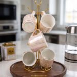 A set of 6 iridescent ceramic tea and coffee mugs hanging on a gold-tone metal stand resting on a wooden tray with a kitchen interior background.