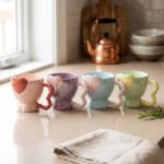 Four colorful ceramic mugs featuring heart accents and beaded handles lined up on a kitchen counter.