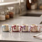 Four pastel ceramic coffee mugs with cute 3D bows and heart-shaped handles lined up on a kitchen counter.