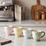 Four pastel ceramic coffee mugs with cute raised bows and matching colorful bases arranged on a marble kitchen counter.