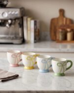 Four pastel ceramic coffee mugs with cute raised bows and matching colorful bases arranged on a marble kitchen counter.